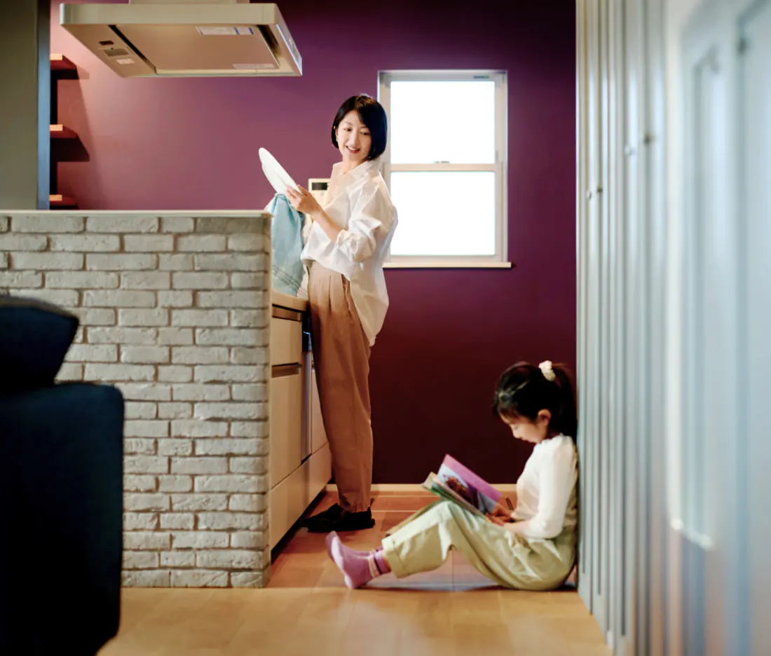 キッチンで皿を片付ける白いシャツ姿の女性と、壁際の床に座って絵本を読む幼い女の子。